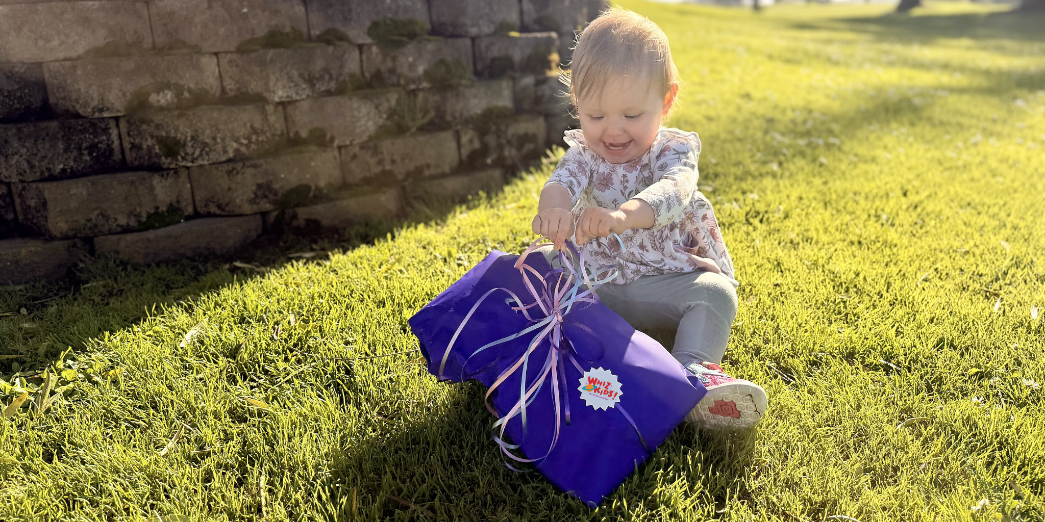 A young child sitting on green grass outdoors, holding a wrapped purple gift box tied with a white ribbon and bow.
